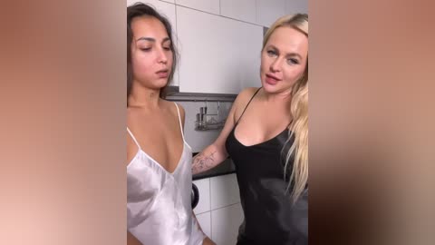Video of two women in a modern bathroom. One with medium skin tone, long dark hair, wearing a white satin robe. The other with fair skin, long blonde hair, in a black satin dress.