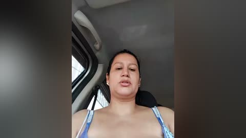 Video of a Latina woman with medium skin tone, dark hair tied back, wearing a blue tank top, sitting in a car, looking forward, with a neutral expression.