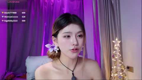 Video of an Asian woman with fair skin, straight brown hair in a ponytail with a purple flower, wearing a black necklace, in a dimly-lit bedroom with purple curtains, white bed, and fairy lights.