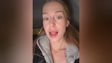Video of a blonde woman with green eyes, wearing a black jacket, open to reveal a gray hoodie, mouth open in a surprised expression, blurred background.
