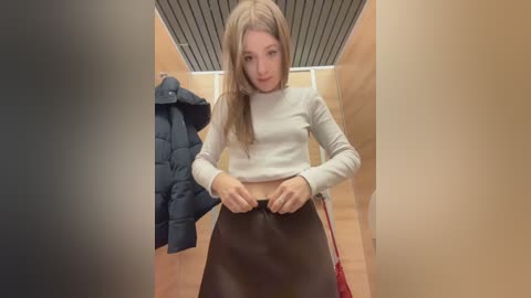A video of a young Caucasian woman with long blonde hair, wearing a beige sweater and a black leather skirt, adjusting the zipper in a changing room with wooden paneling and a dark blue jacket hanging on a hook.