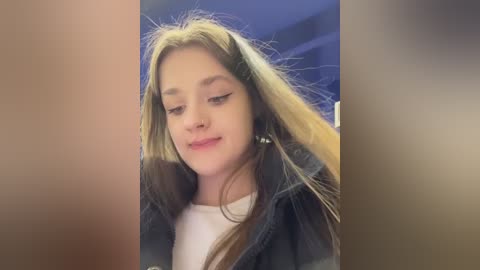 A video of a young girl with long blonde hair, wearing a denim jacket, sitting indoors with a blurred background.