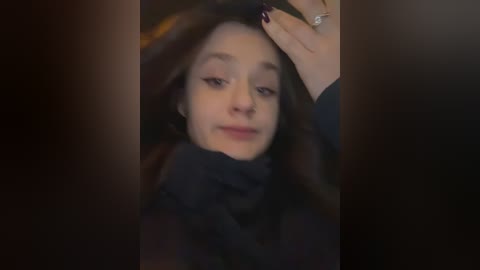 A video of a young woman with fair skin, dark hair, and wearing a black coat, adjusting her hair with one hand while the other rests on her head. The background is blurry, with a dim, warm light.