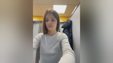 Video of a young Caucasian woman with straight, shoulder-length brown hair, wearing a light grey long-sleeve shirt, standing in a public restroom with tiled walls and a ceiling light.