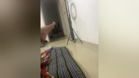 A video of a dimly lit, sparse room with a beige floor. A person, possibly male, wearing shorts and a red shirt, sits on a black tripod facing a mirror. A colorful, patterned blanket lies nearby.