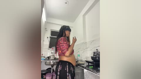 A video shows a woman with long black hair, wearing a red and white patterned crop top and black pants, cooking in a small, cluttered kitchen with white walls and marble countertops.