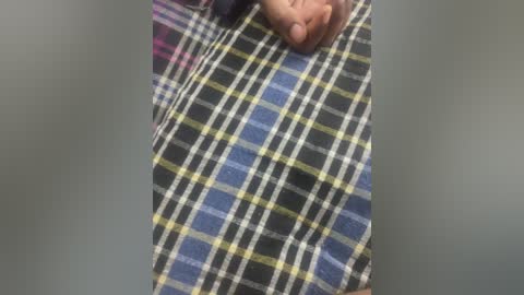 A close-up video of a person's hand, partially visible, resting on a blue, yellow, and black plaid fabric, likely a shirt. The background is blurred, focusing on the hand and fabric texture.