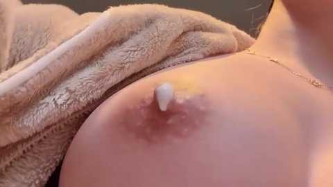 Video of a woman's bare breast with a visible nipple, partially covered by a soft, beige, fuzzy blanket. The background is blurred, suggesting an intimate indoor setting.