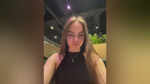 Video of a young Caucasian woman with long brown hair, wearing glasses and a black sleeveless top, standing in a modern indoor setting with green plants and industrial ceiling lights.