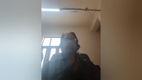 A video of a middle-aged man with glasses, a beard, and dark clothing, standing in a dimly lit room with beige walls and a window to the left.