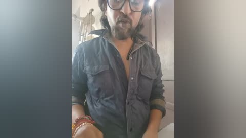 Video of a man with medium brown skin, wearing glasses, a denim shirt, and a bracelet, standing in a dimly lit room with a statue in the background.