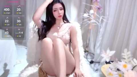 Video of an East Asian woman with long black hair, fair skin, and large breasts, sitting on a white fur rug in a room with white curtains, flowers, and a digital display showing her stats.
