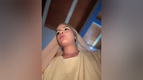 Video of a Caucasian woman with light skin, blonde hair, wearing a beige T-shirt, looking up while adjusting her hair, in a room with wooden beams and blue accents.