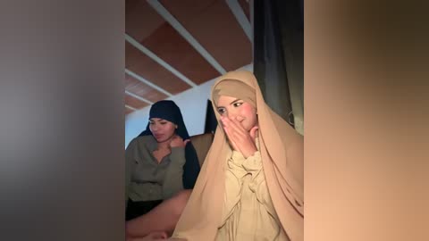 Video of two women in traditional Muslim attire, one with a headscarf and long beige dress, the other in a green top, sitting indoors with a tiled ceiling.
