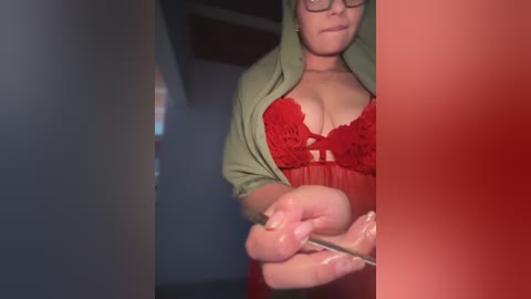 Video of a fair-skinned woman with glasses, wearing a red lace bra and olive-green robe, holding a knife, in a dimly lit room.