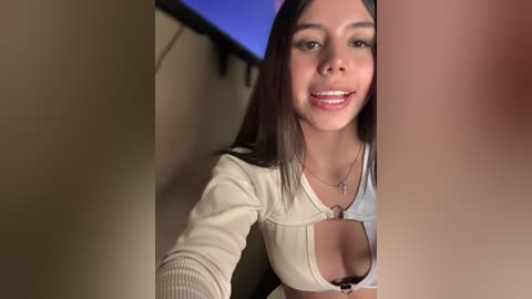Video of a young Latina woman with long dark hair, smiling, wearing a low-cut white top that reveals cleavage. She's indoors, in a dimly lit room with a blue light in the background.
