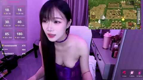 A video of a young East Asian woman with long black hair, wearing a purple strapless top, sitting at a desk in a dimly lit room, with a Minecraft game open on her screen.