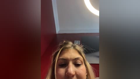 A video of a blonde woman with fair skin, slightly blurred, standing in a red-walled room with a circular light fixture on the ceiling.
