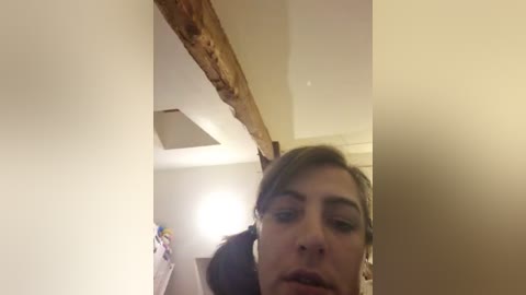 Video of a woman with light skin and straight brown hair, partially visible, in a bathroom with a wooden beam, white walls, and a towel hanging.