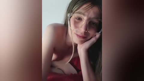 Video of a topless Caucasian woman with long brown hair and glasses, resting on a red bed, with a blurred background.