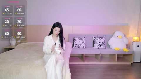 A video of an East Asian woman in white bathrobe, sitting on a bed in a minimalist bedroom with a plush duck, white bedding, and a nightstand.