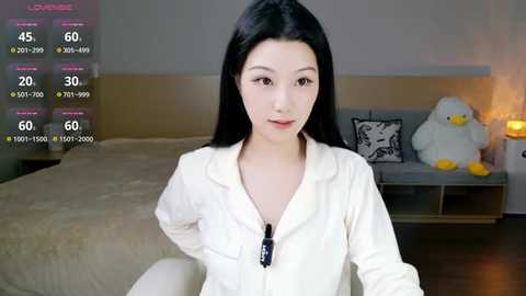 A video of an Asian woman with long black hair, wearing a white zip-up hoodie, standing in a modern bedroom with a bed, white duck, and nightstand with a lamp.