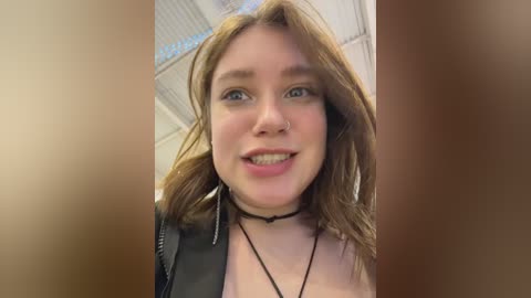 Video of a young woman with fair skin, light brown hair, and blue eyes, wearing a black choker and black jacket, smiling, taken indoors with ceiling lights visible in the blurred background.