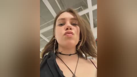 Video of a young woman with light skin, medium-length brown hair, wearing a black choker necklace and dangling earrings, captured from a low angle in a modern indoor setting with a white ceiling.