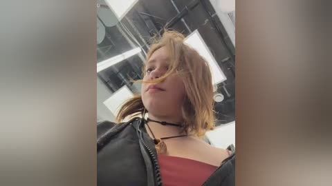 Video of a young woman with messy blonde hair, wearing a black leather jacket and a red top, standing in a modern, dimly lit room with metallic accents.