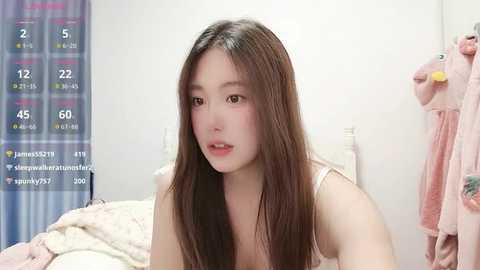A video of an East Asian woman with long brown hair, wearing a white tank top, sitting on a bed in a pink room. A calendar with dates and a phone are visible in the background.