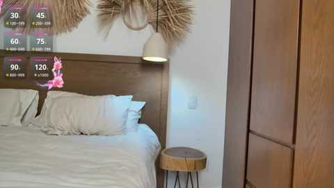 Video of a minimalist bedroom with a white bed, wooden headboard, beige pendant light, and a round wooden side table.