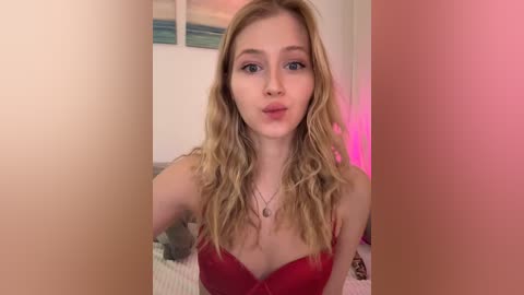 Video of a young, fair-skinned woman with long, wavy blonde hair and blue eyes, wearing a red lace bra, posing indoors with a pinkish glow.