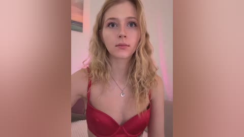 Video of a fair-skinned, blonde woman with wavy hair, wearing a red bra, standing in front of a blurred pink background.