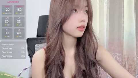 Video of a young Asian woman with long, wavy brown hair, wearing a sleeveless top, sitting in a gaming chair in a well-lit room. Background includes a monitor displaying game stats and a window with sheer curtains.