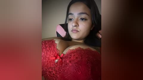 Video of a young woman with medium skin tone, straight black hair, and wearing a red lace bra with a bow, lying on her side with a pink phone, blurred background.