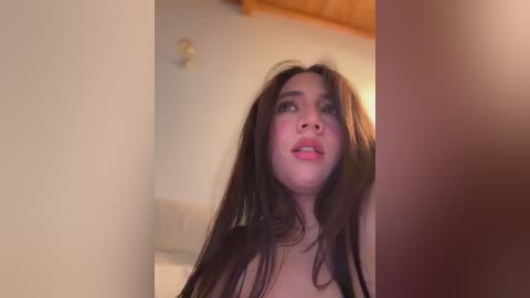 A video of a young woman with long, dark hair, fair skin, and full lips, looking upward with a slight smile, in a dimly lit room with wooden ceiling beams.