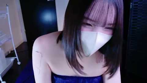 Video of an Asian woman with straight, shoulder-length black hair, wearing a white face mask and a blue velvet dress. She is indoors, standing in front of a black wall, with a white metal stand in the background.