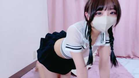 A video of an East Asian woman in a white sailor-style crop top, black skirt, and face mask, on all fours on a pink carpet, with a white wall and pink curtain background.