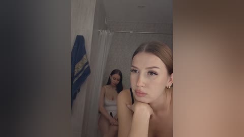 Video of a woman with fair skin and straight brown hair, in a white towel, looking contemplative in a dimly lit bathroom. Another woman, also fair-skinned, with long brown hair, wearing a blue towel, stands in the background.