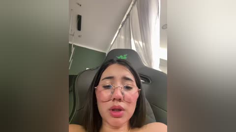 A video shows an Asian woman with glasses, fair skin, and pink lipstick, sitting in a green chair with a green curtain behind her.