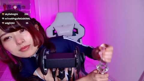 Video of an Asian woman with long brown hair, wearing a navy blue top, sitting in a pink-lit room. She's holding a microphone and a glass, with a gaming chair and a computer screen in the background.