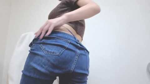 A video of a person's lower back and buttocks, wearing tight, blue jeans with a white bra visible, in a white-walled bathroom.