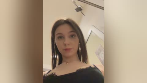 Video of a young woman with light skin, dark brown hair, wearing a black lace camisole. She has a slim physique, small breasts, and a neutral expression. Background includes a beige wall and a ceiling light.