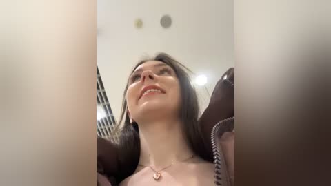 A video captures a smiling woman with long dark hair, wearing a brown jacket, in a blurred indoor setting.