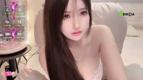 Video of a young East Asian woman with long black hair, pale skin, and a slender figure, wearing a pink lace bra, sitting on a beige couch.