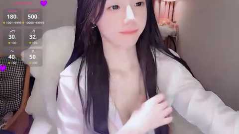 Video of an East Asian woman with long black hair, fair skin, and a white blouse, capturing a selfie with her face partially covered by a Band-Aid. Background features a bed and a checkered-patterned shirt.