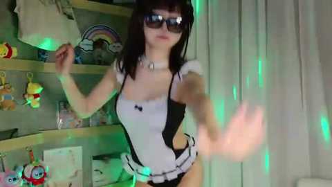 Video of a slender, pale-skinned woman in a revealing black and white maid costume, with large sunglasses, standing in a dimly lit room filled with stuffed toys and green glow sticks.