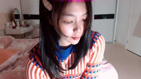 Video of an East Asian woman with long black hair wearing a purple headband, striped shirt, and blue collar, sitting in a softly lit bedroom with pink sheets, white walls, and a vanity table.
