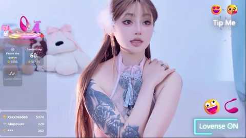 A video of an East Asian woman with long brown hair, light skin, and a sleeve tattoo, wearing a pastel pink top, in a room with a teddy bear and a blue background.