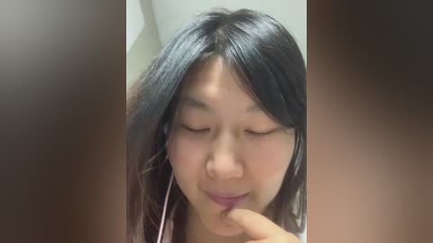 Video of an Asian woman with long black hair, wearing headphones, gently touching her lips with her finger, smiling softly, with blurred faces in the background.